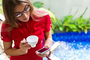 Affluent Prints: Woman reading Mails on smartphone while drinking coffee on balcony in tropical setting