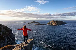 Newfoundland Prints: Woman in red jacket is standing at the edge of a cliff with open arms and enjoying the beautiful ocean scenery