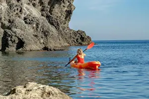 Kayaking Prints: A woman in a red swimsuit paddles an orange kayak in the calm blue sea