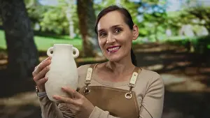 Pleasant Prints: Woman smiling outdoors in a green park holding a white vase wearing a brown apron showcasing artistry