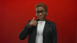 Worry Prints: Woman wearing glasses and hoop earrings in black blazer and white top points thumb to chest in red studio; doubt