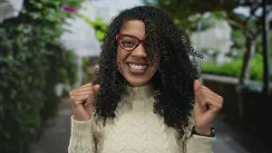 Thrill Prints: Woman wearing red glasses and cream sweater clenches fists with broad smile in green park; celebration joy