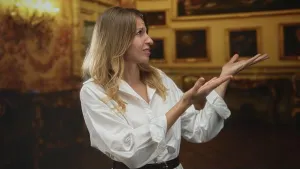 Concern Prints: Woman in white blouse gesturing with open palms raised amid framed paintings in ornate building gallery; art discussion