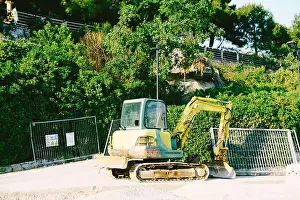Scoop Prints: Yellow crawler tractor with a bucket working in the park against the background of a metal fence