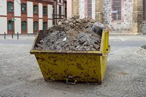 Rubble Prints: A yellow metal container filled with construction debris stands in a square in front of historic buildings