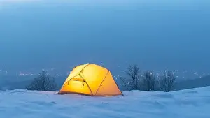 Yellow tent lighted from the inside against the backdrop of glowing city lights in fog. Amazing snowy landscape. Tourists camp in winter mountains
