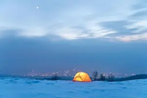 Yellow tent lighted from the inside against the backdrop of glowing city lights in fog. Amazing snowy landscape. Tourists camp in winter mountains