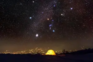 Yellow tent lighted from the inside against the backdrop of glowing city lights and incredible starry sky. Amazing night landscape