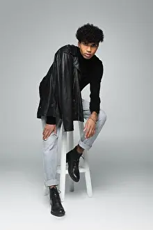 Stool Prints: young african american man with black leather jacket on shoulder posing on high stool on grey background