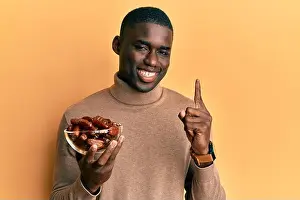 Opinion Prints: Young african american man holding bowl with dates smiling with an idea or question pointing finger with happy face