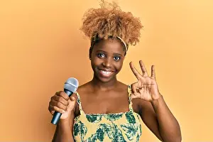 Approval Prints: Young african woman with afro hair singing song using microphone doing ok sign with fingers