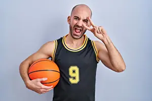 Shaved Head Prints: Young bald man with beard wearing basketball uniform holding ball doing peace symbol with fingers over face
