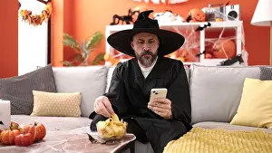 Texting Prints: Young bald man wearing wizard costume eating chips potatoes using smartphone at home