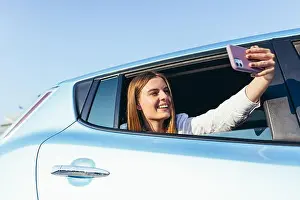 Video Call Prints: Young beautiful woman talking on video call sitting in the car sticking his hand with the phone out the window