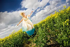 Airy Prints: young blonde woman in grecian style cream teal dress jumping floating above field of yellow rapeseed with blue sky