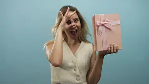 Wrap Prints: Young blonde woman holding pink gift box and making ok gesture with right hand in blue studio; playful celebration