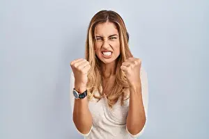 Young blonde woman standing over isolated background angry and mad raising fists frustrated