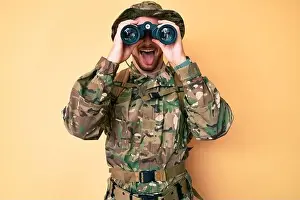 Sticking Prints: Young caucasian man wearing camouflage army uniform using binoculars sticking tongue out happy with funny expression