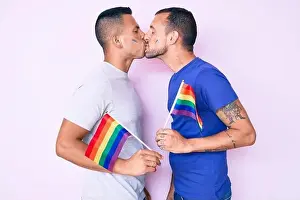 Lgbt Prints: Young couple of gay men together in love holding homosexual pride colorful flag kissing with affection
