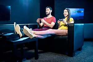 Gamer Prints: Young couple playing video games with gaming console sitting on the couch in the dark room of the playing club