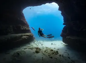 Tropical Island Prints: A young female free diving down into a cave doorway accompanied by a green sea turtle with clear blue water