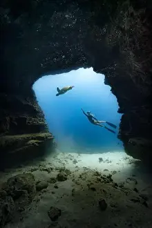 Tropical Island Prints: A young female free diving down into a cave doorway accompanied by a green sea turtle w/clear blue water