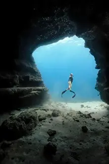 Tropical Island Prints: A young female free diving down into a cave doorway in the clear blue waters of Hawaii framed by shadowy cave walls