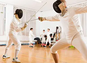 Young fencers looking at fencing duel with foils of two professional trainers in gym