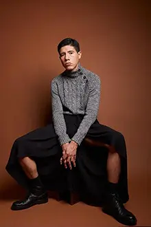 Hands Clasped Prints: Young gay in boots and trendy wear sitting with clasped hands while looking at camera