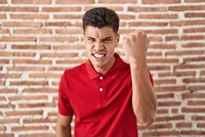Punching Prints: Young hispanic man standing over bricks wall angry and mad raising fist frustrated