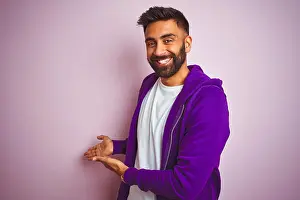 Optimistic Prints: Young indian man wearing purple sweatshirt standing over isolated pink background Inviting to enter smiling natural