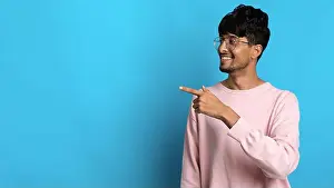 Poses Prints: Young Man in Pink Sweater Smiles and Points at Blue Background in Studio Setting