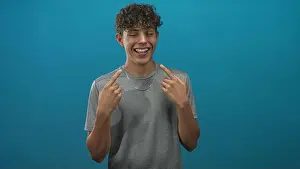 Dental Prints: Young man pointing fingers at his teeth and smiling broadly in blue studio backdrop; joy youth energy