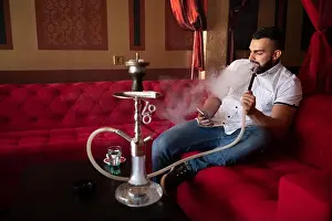 Young Man Smoking Shisha At Arabic Restaurant - Man Exhaling Smoke Inhaling From A Hookah and Texting on Phone