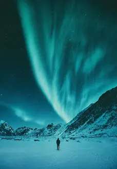 Young man is standing on a remote beach watching the Northern Lights dance above mountains in winter