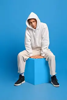 Pullover Prints: Young man in white sweatshirt with hood and sweatpants sits on wooden cube over blue studio background