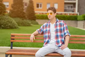 One Alone Prints: Young modern looking fashionable man posing by sitting on bench in the park