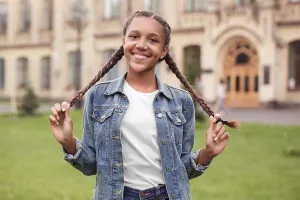 Descent Prints: Young mulatto girl wearing jeans jacket standing at school yard holding braids looking camera smiling playful