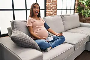 Young pregnant woman using blood pressure monitor sitting on the sofa angry and mad screaming frustrated and furious