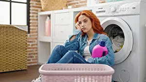 Redhead Prints: Young redhead woman holding detergent bottle leaning on washing machine tired at laundry room