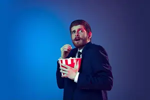 Shocked Prints: Young scared man in business suit eating popcorn and watching movie isolated on dark blue background