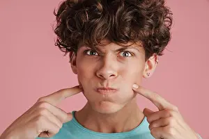Cheek Prints: Young teenager with curly hair blowing his cheeks isolated over pink background