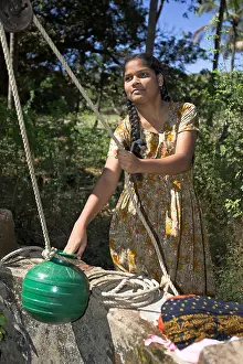Fashioned Prints: Young woman draws water from well by hand with green pot on traditional rope pulley Mandrem village Goa