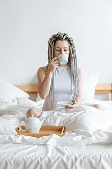 Generation X Prints: Young woman drinking tea from cup sitting in white bed