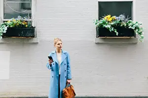 Purse Prints: Young woman holding smart phone and purse while standing against building in city