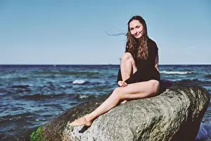 Barefoot Prints: Young woman with long brown hair in black outfit sits barefoot on large rock at Baltic Sea shoreline