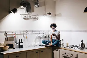 Cross Legged Prints: Young woman sitting on kitchen worktop, checking messages on smartphone