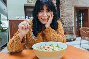 Housewife Prints: young woman sitting leaning from table with one hand on her face surprised happy smiling with fork and bowl of salad