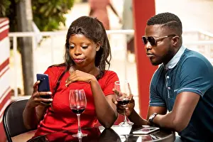 Wine Glass Prints: young woman sitting outside a refreshment booth watching cell phone while her boyfriend looks at her wineglass
