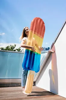 Inflatable Prints: Young woman standing barefoot on balcony with an ice lolly shaped airbed